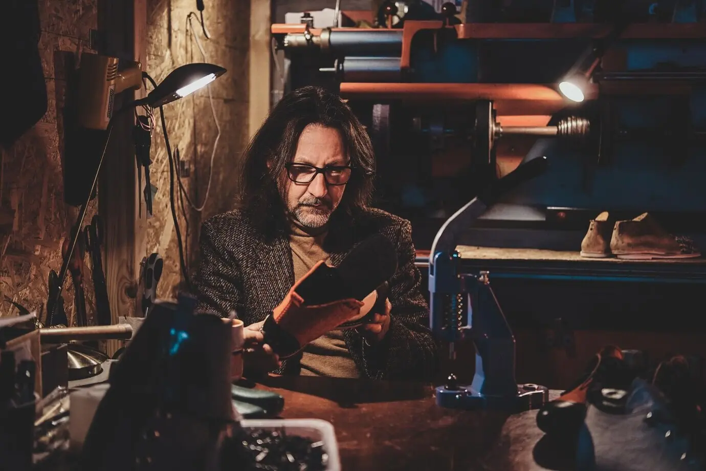 A thoughtful shoemaker wearing glasses is working on his new shoe collection at his workshop.
